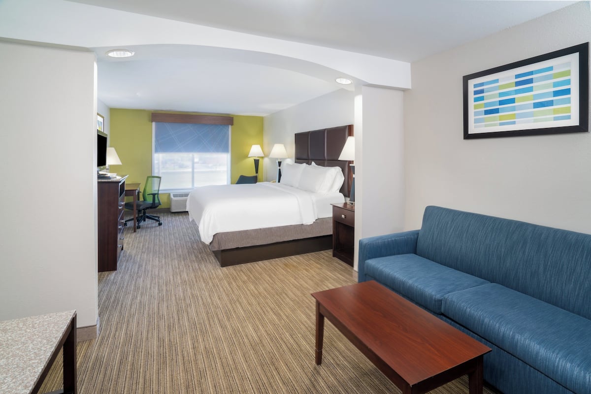 A hotel room with a bed, a blue sofa, a wooden table, a desk with a chair, and a window with a view.