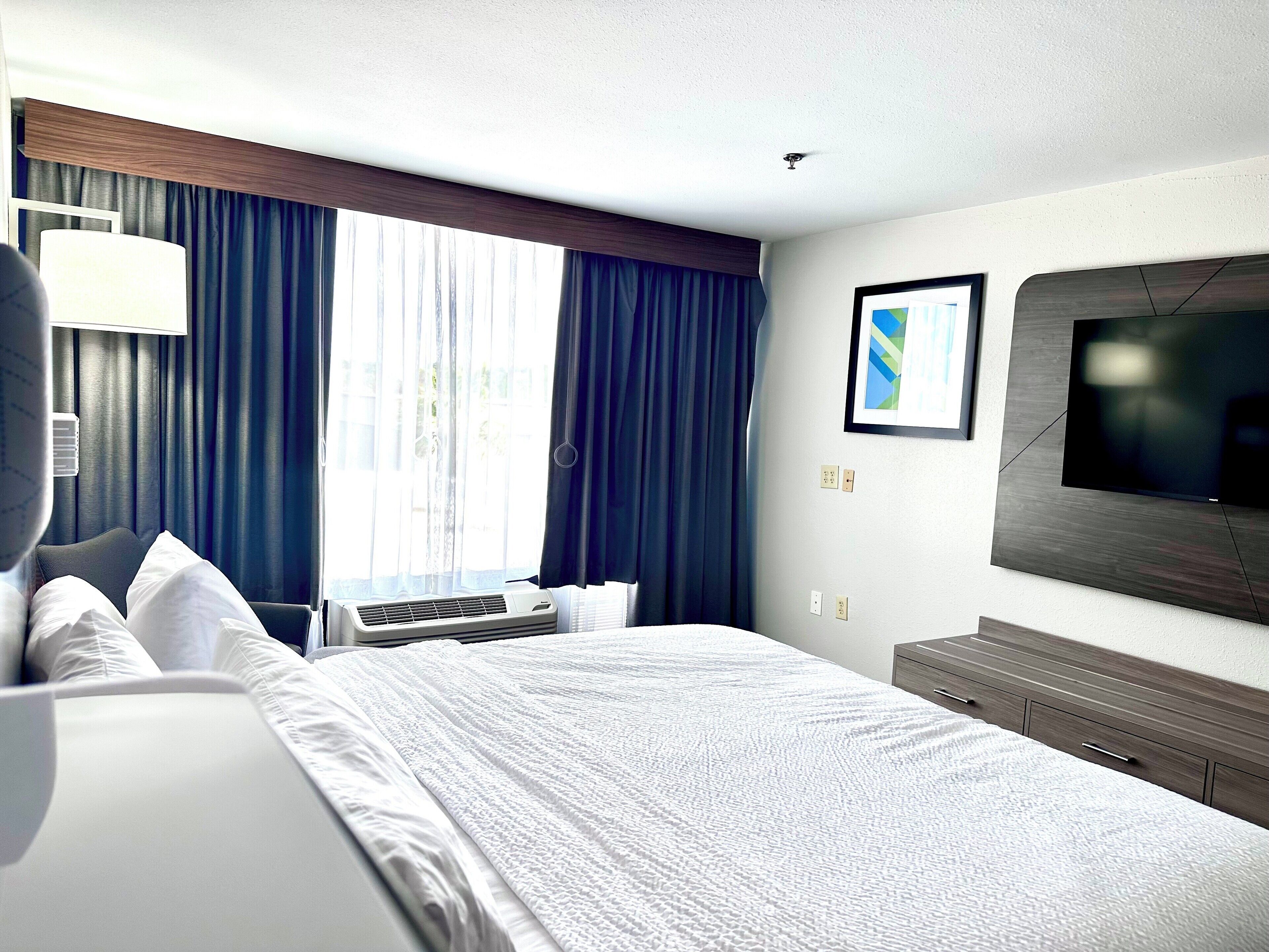 A hotel room with a large bed, a flat-screen TV mounted on the wall, a window with curtains, and a bedside table.
