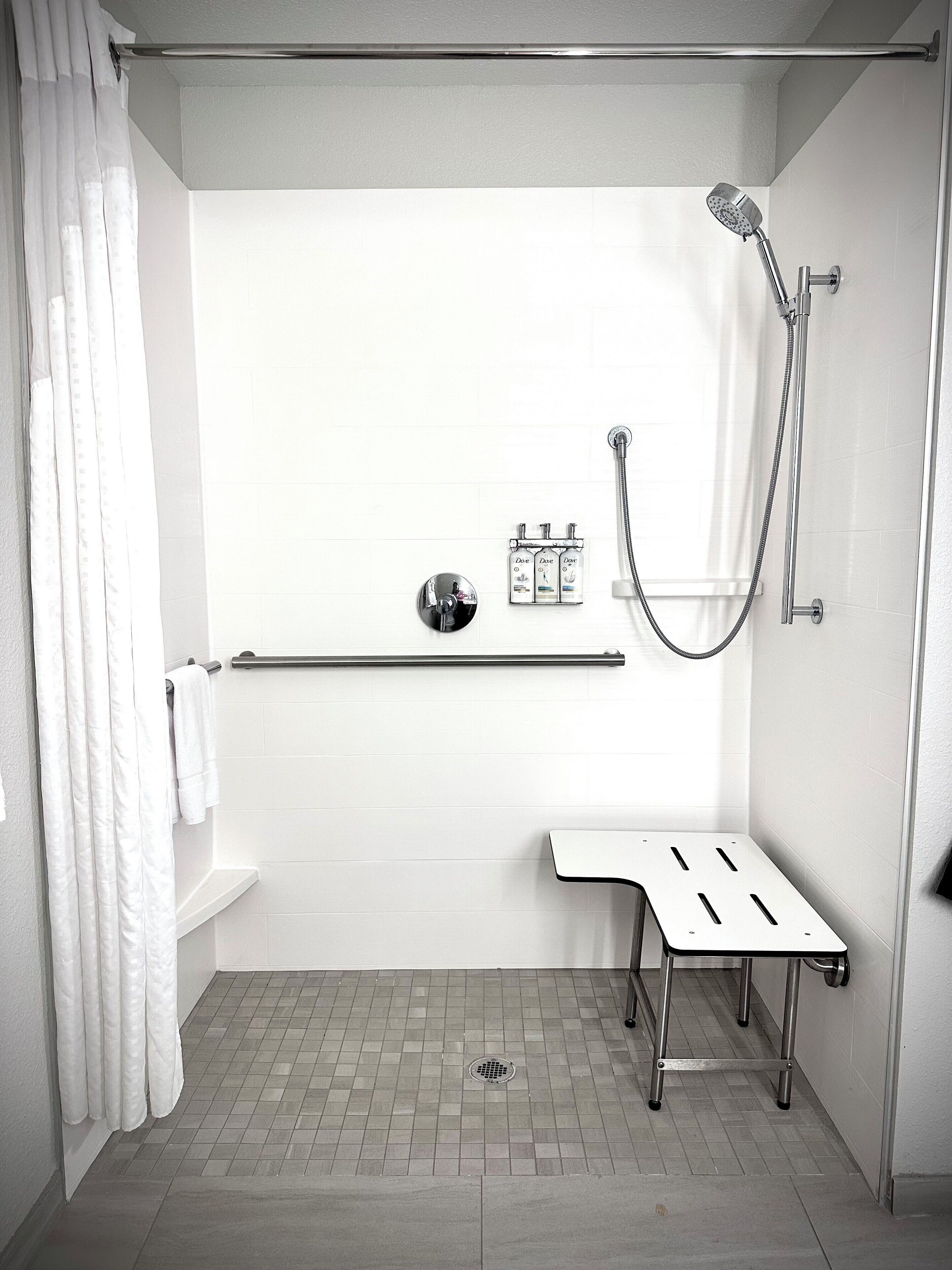 A modern shower area with a handheld showerhead, a handheld shower attachment, and a wall-mounted faucet. There is a towel rack with a towel hanging on it, and a small shelf with toiletries.