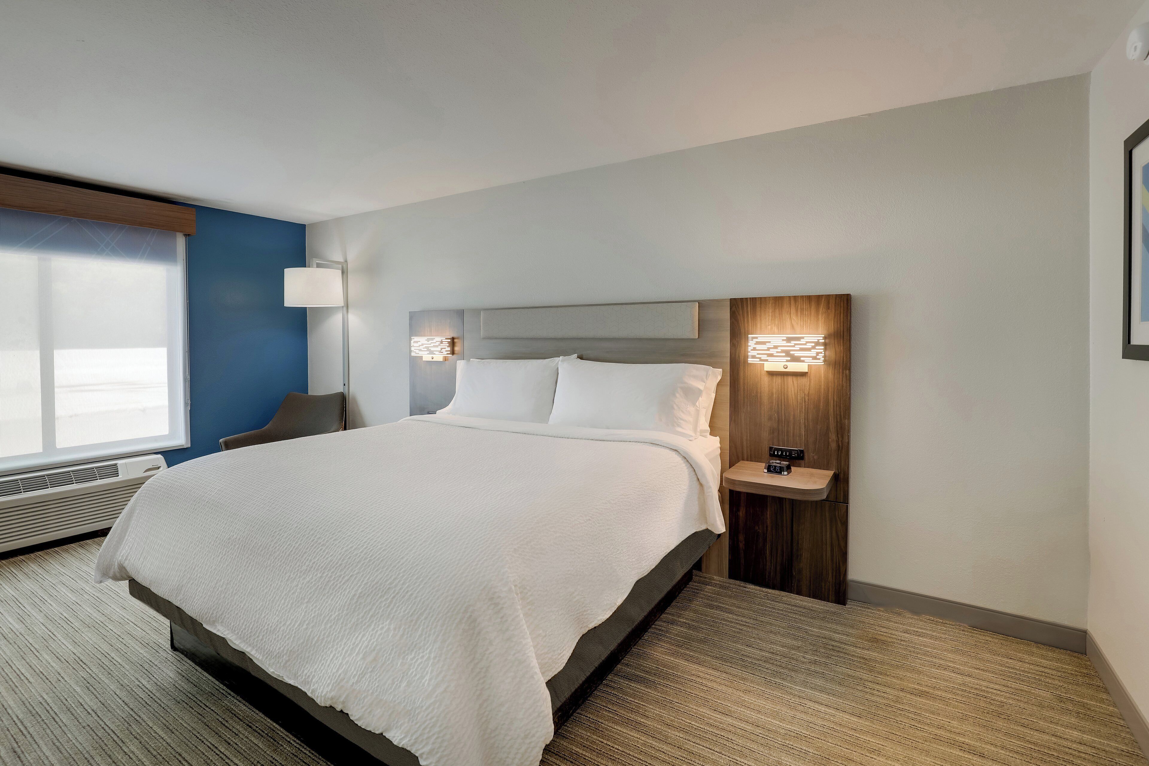 Una habitación de hotel moderna con una cama grande, una mesita de noche con lámpara y una ventana con persianas.