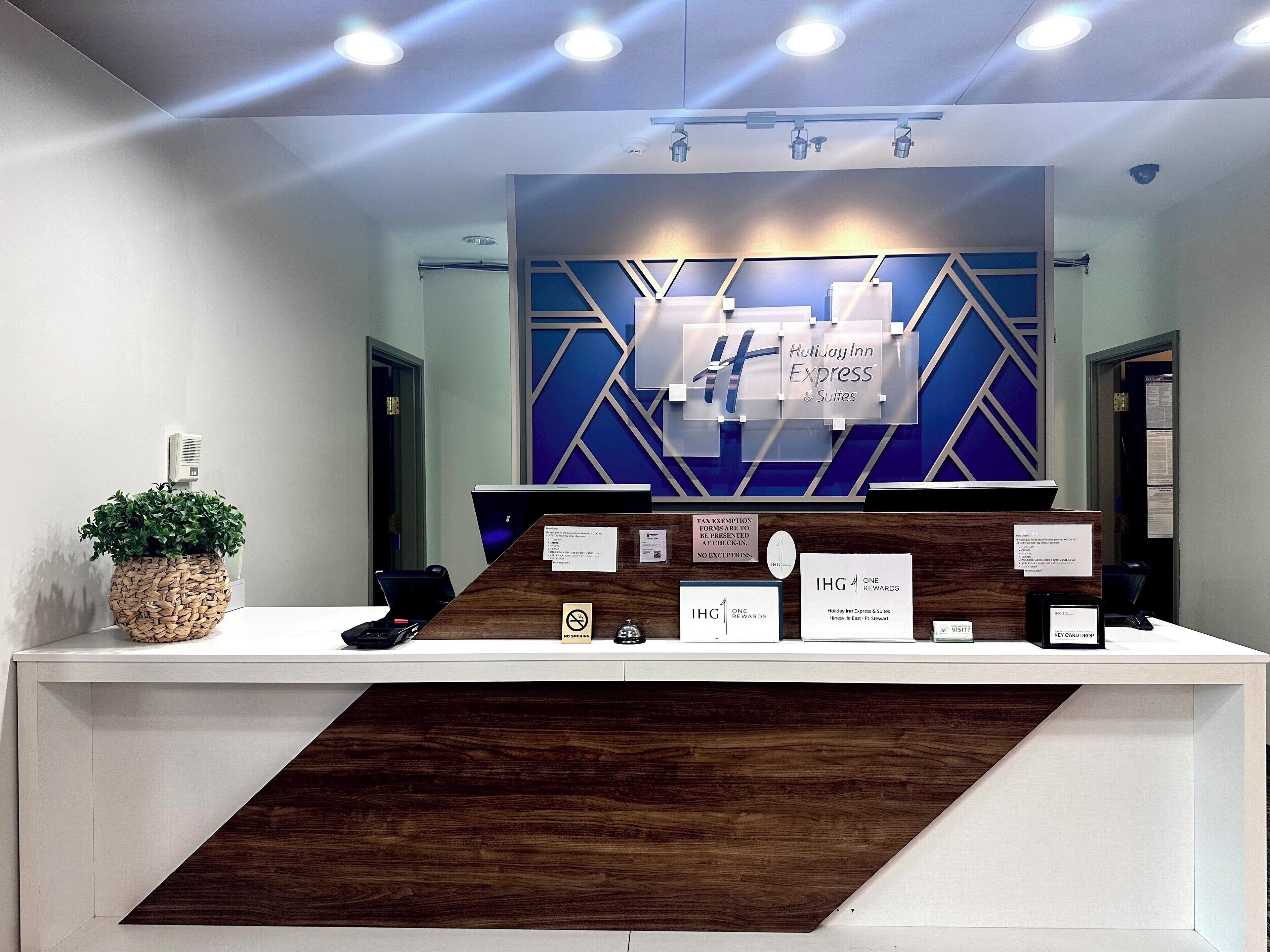 A hotel reception desk with a modern design, including a digital display, a plant, and informational signs about hotel amenities and policies.