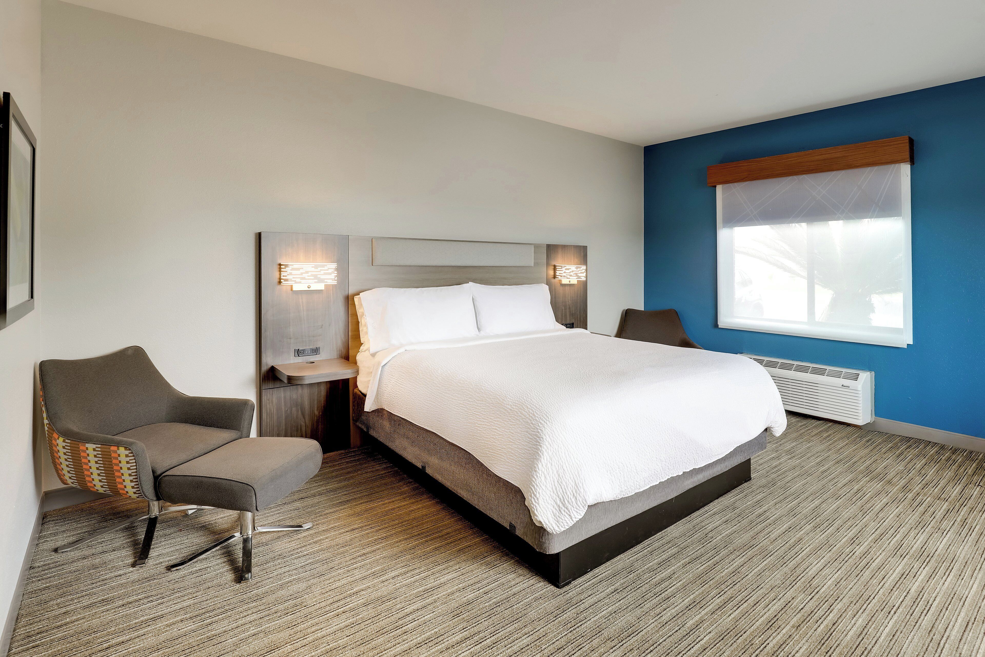 A hotel room with a large bed, a chair, a nightstand, and a window with blinds.
