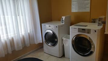 Una sala de lavandería con dos lavadoras de carga frontal, una mesita con una cafetera y un panel de control en la pared.