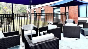 A patio area with outdoor furniture and umbrellas, a brick building in the background, and a metal fence. - Holiday Inn Express Hinesville by IHG (Hinesville)