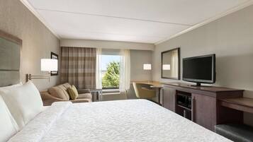 A hotel room with a large bed, a desk with a flat-screen TV, a chair, and a window with curtains.