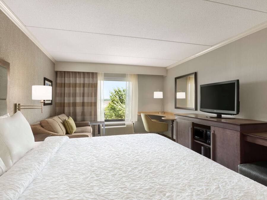A hotel room with a large bed, a desk with a flat-screen TV, a chair, and a window with curtains.