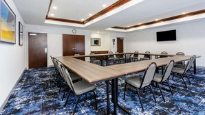 Een congreszaal met een lange tafel en stoelen, houten plafondpanelen en een blauw kamerbreed tapijt met dessin.