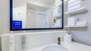 Uma casa de banho com uma pia branca, um espelho e prateleiras com toalhas dobradas. Existem duas embalagens de sabonete líquido e uma caixa de lenços na bancada.