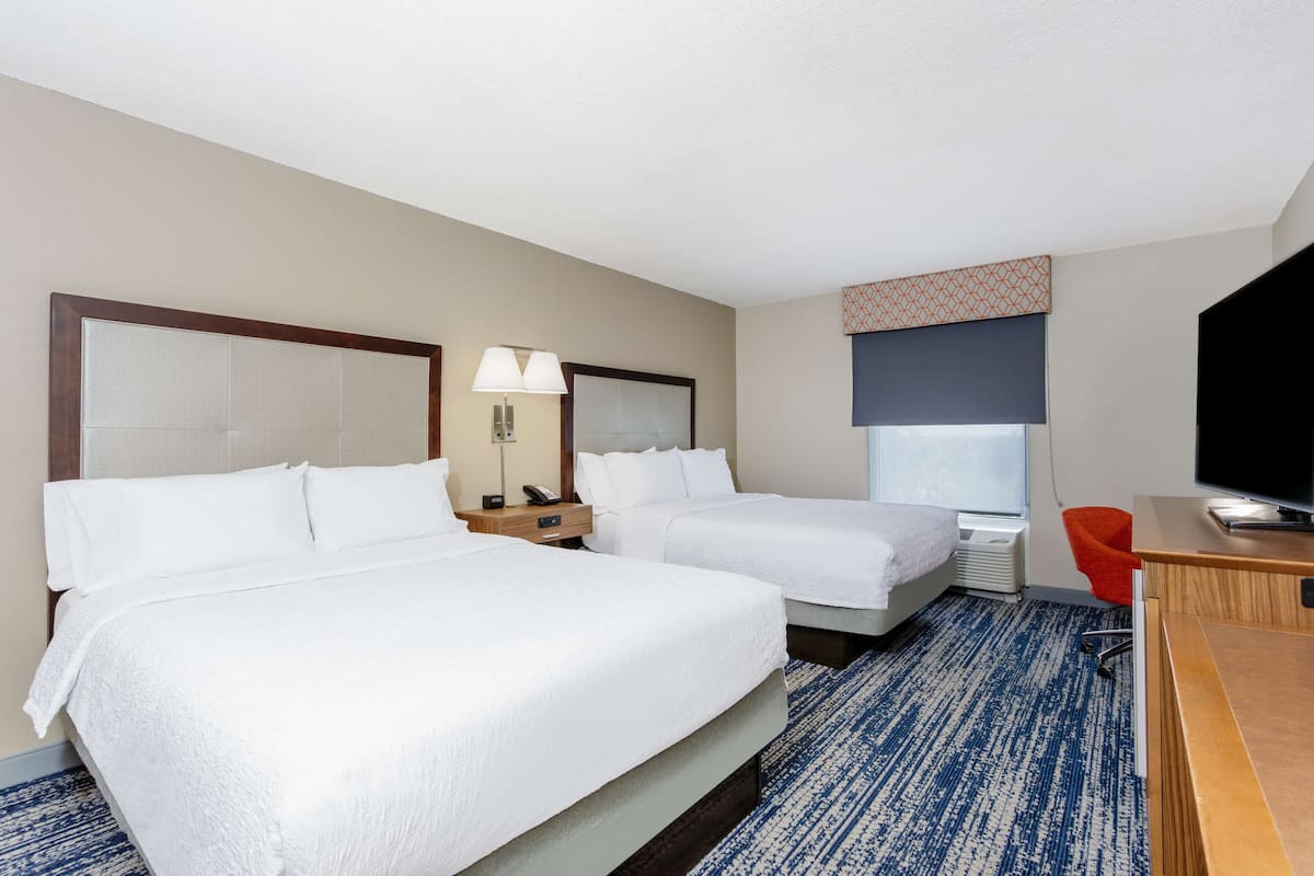 Habitación de hotel con dos camas, un escritorio, una silla, un televisor y una ventana con persiana estampada.