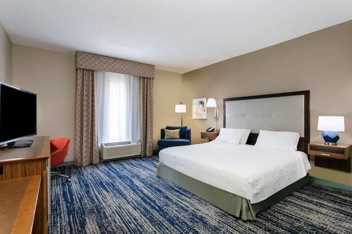 Habitación de hotel con una cama grande, televisión, escritorio, silla y ventana con cortinas.