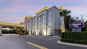 Een moderne hotelentree met een bord met de tekst 'Welcome', een 'Hampton Inn by Hilton' bord en een parkeerplaats.