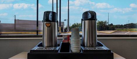 A coffee machine with a cup holder, a can of coffee, and a stack of paper cups on a table.