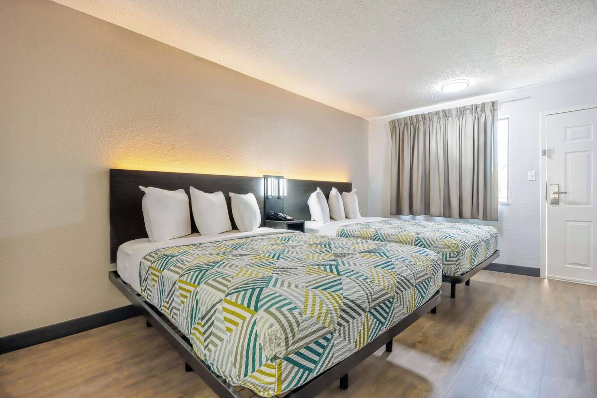 A hotel room with two beds, a headboard with a light fixture, a bedside table, and a window with curtains.