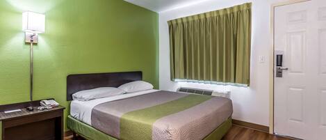 A hotel room with a bed, a bedside table with a lamp, a window with curtains, and a door.