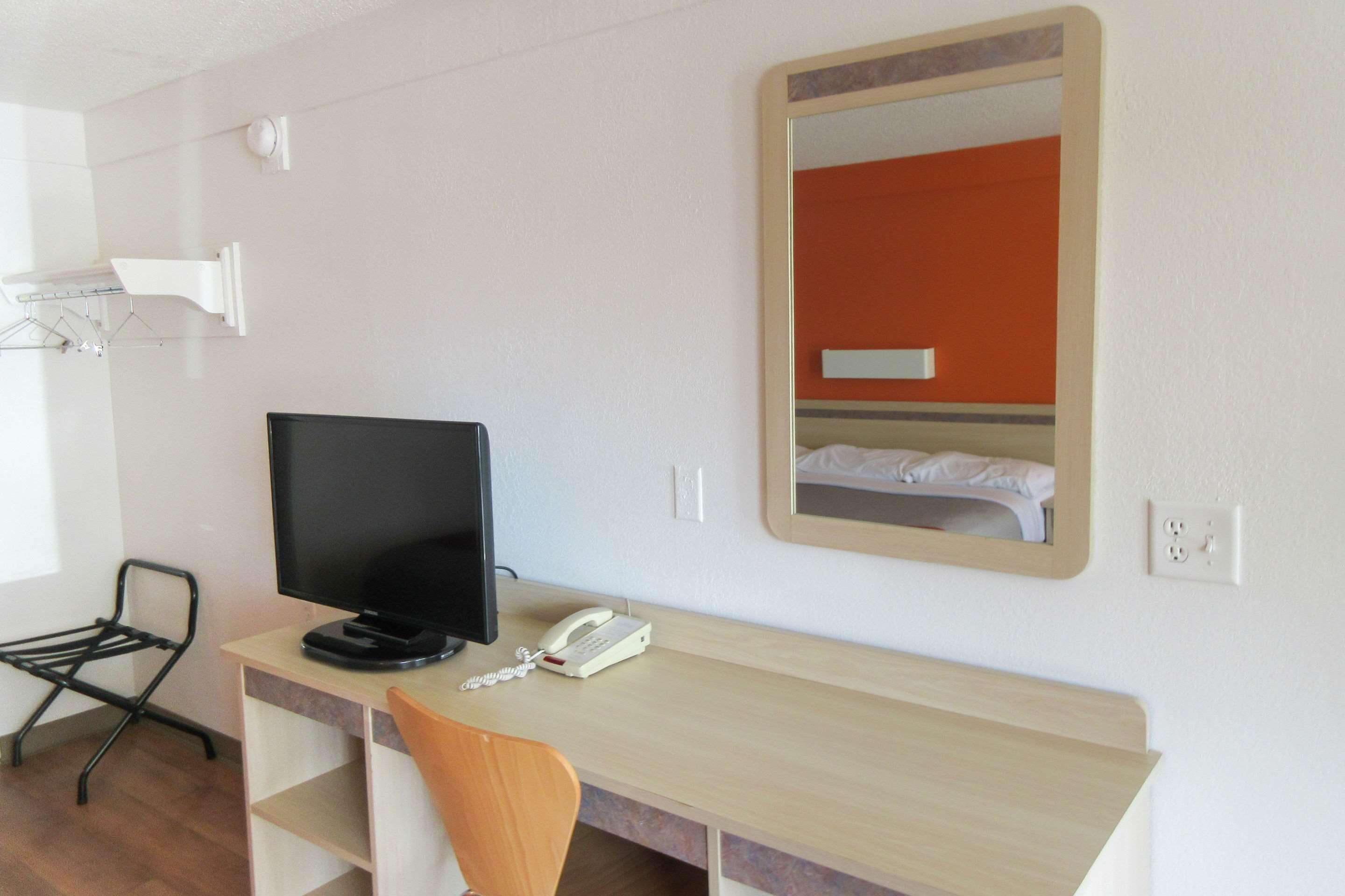 A hotel room with a wooden desk, a black TV, a white telephone, and a mirror reflecting a bed with white bedding.