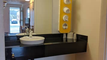 A bathroom with a black countertop, white sink, and a mirror above it. There is a yellow towel rack with three towels and a small window above the sink.