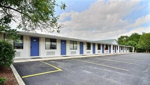 A motel with a row of rooms, each with a blue door and a window. There is a parking lot in front of the building.