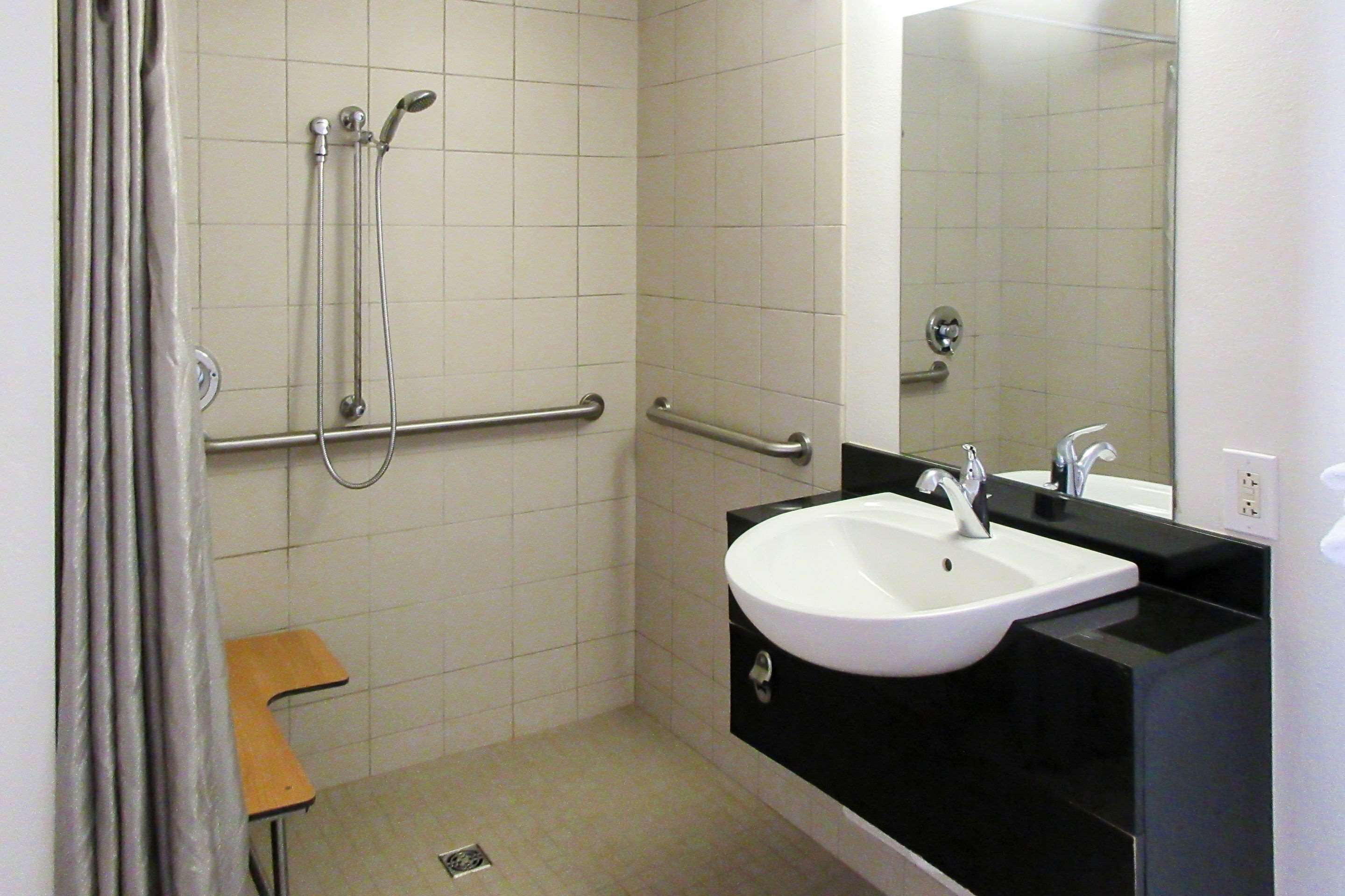 Baño con lavamanos blanco, encimera negra y espejo. Hay barras de apoyo en las paredes y un cabezal de ducha.