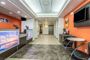 A hotel reception area with a digital screen displaying a message, a reception desk, an elevator, and a small seating area with chairs. - Motel 6 Fort Mill, SC - Charlotte (Fort Mill)