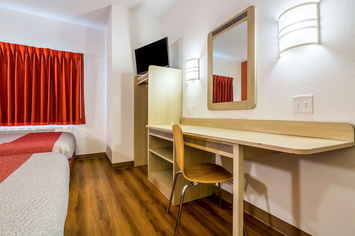 A hotel room with a bed, a desk with a chair, a TV, a mirror, and a window with curtains.