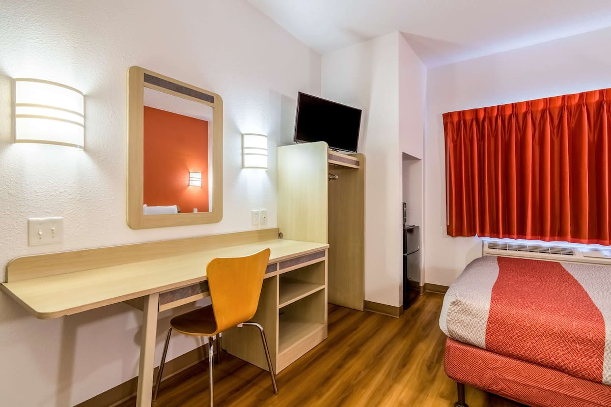 A hotel room with a wooden desk, a chair, a bed with a red and white blanket, a TV on a stand, and a red curtain.