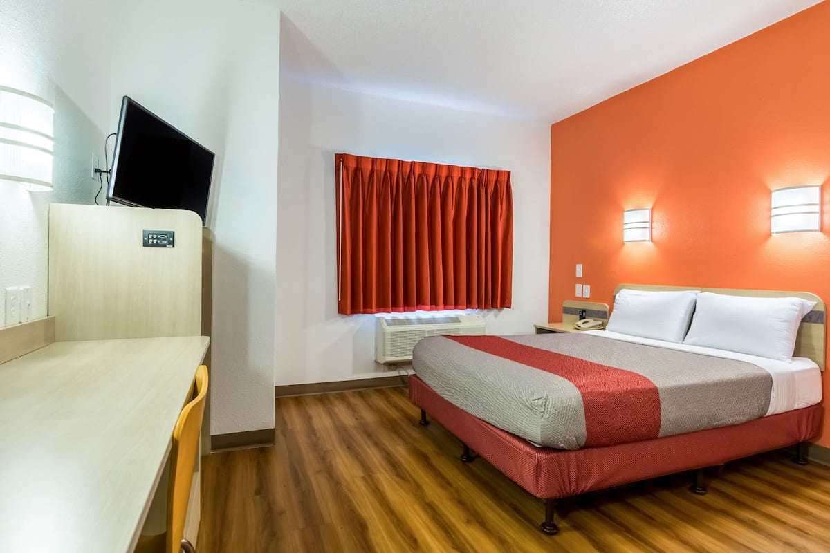 A hotel room with a bed, a desk, a TV, a window with curtains, and a wall-mounted light fixture.