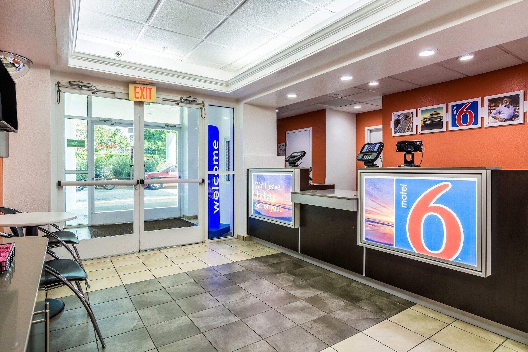 Een hotel receptie met een glazen deur, een digitaal scherm met een bericht en een balie met een bord waarop de naam van het hotel staat.