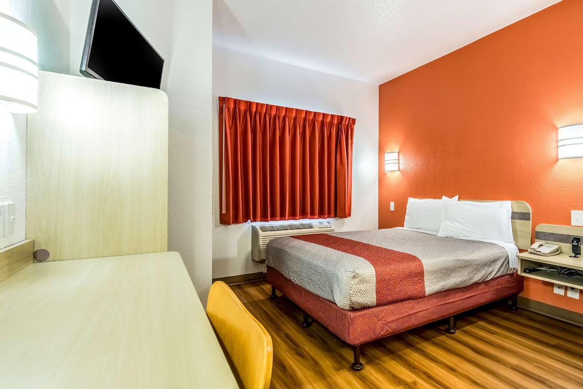 A hotel room with a bed, a desk, a chair, a window with red curtains, and a wall-mounted heater.