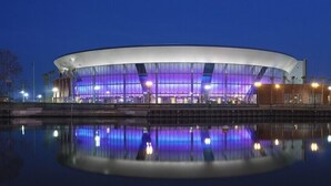 Um edifício moderno e circular, com fachada de vidro e reflexo na água em frente.