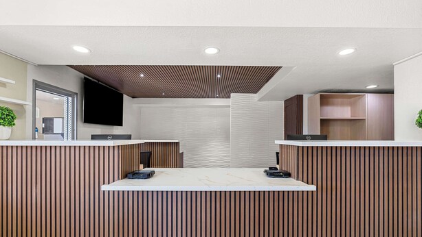 Uma área de recepção com design moderno, destacando um balcão de madeira, uma TV de tela plana na parede e um teto com iluminação embutida.