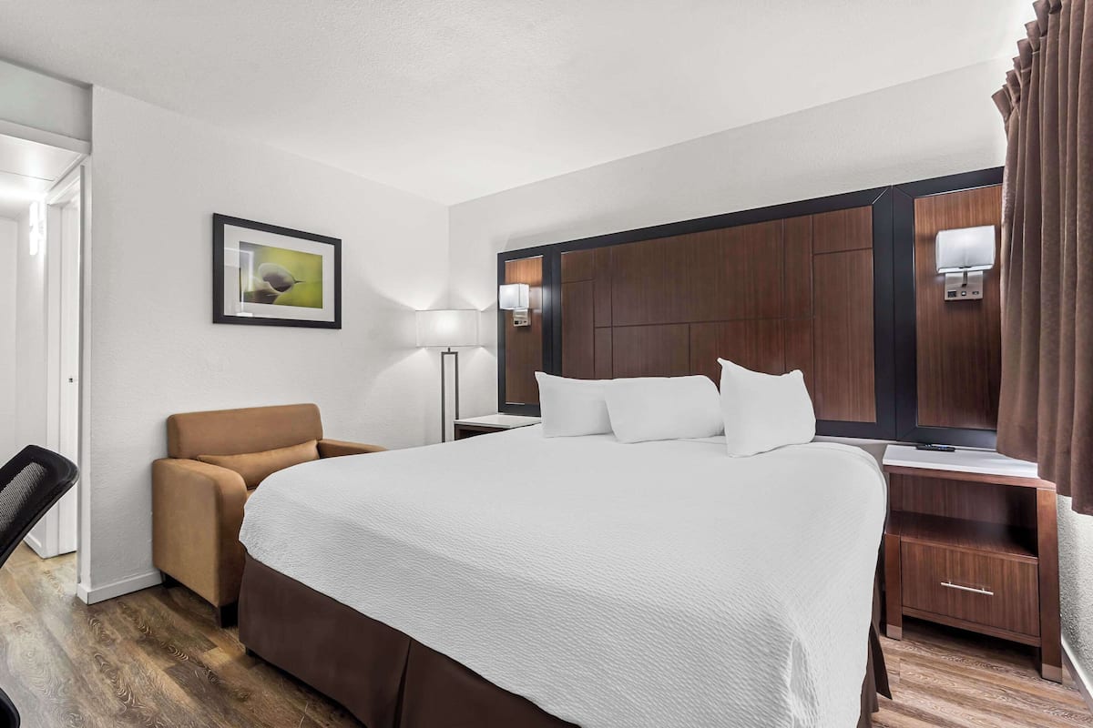 A hotel room with a large bed, a desk with a lamp, a chair, a wardrobe, and a framed picture on the wall.