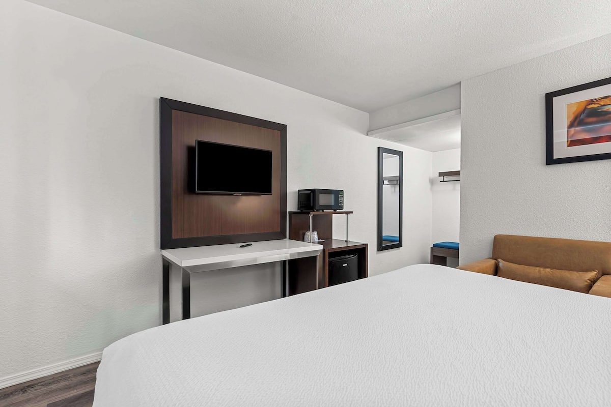 A hotel room with a bed, a TV mounted on a wooden panel, a microwave, a small table, and a chair.