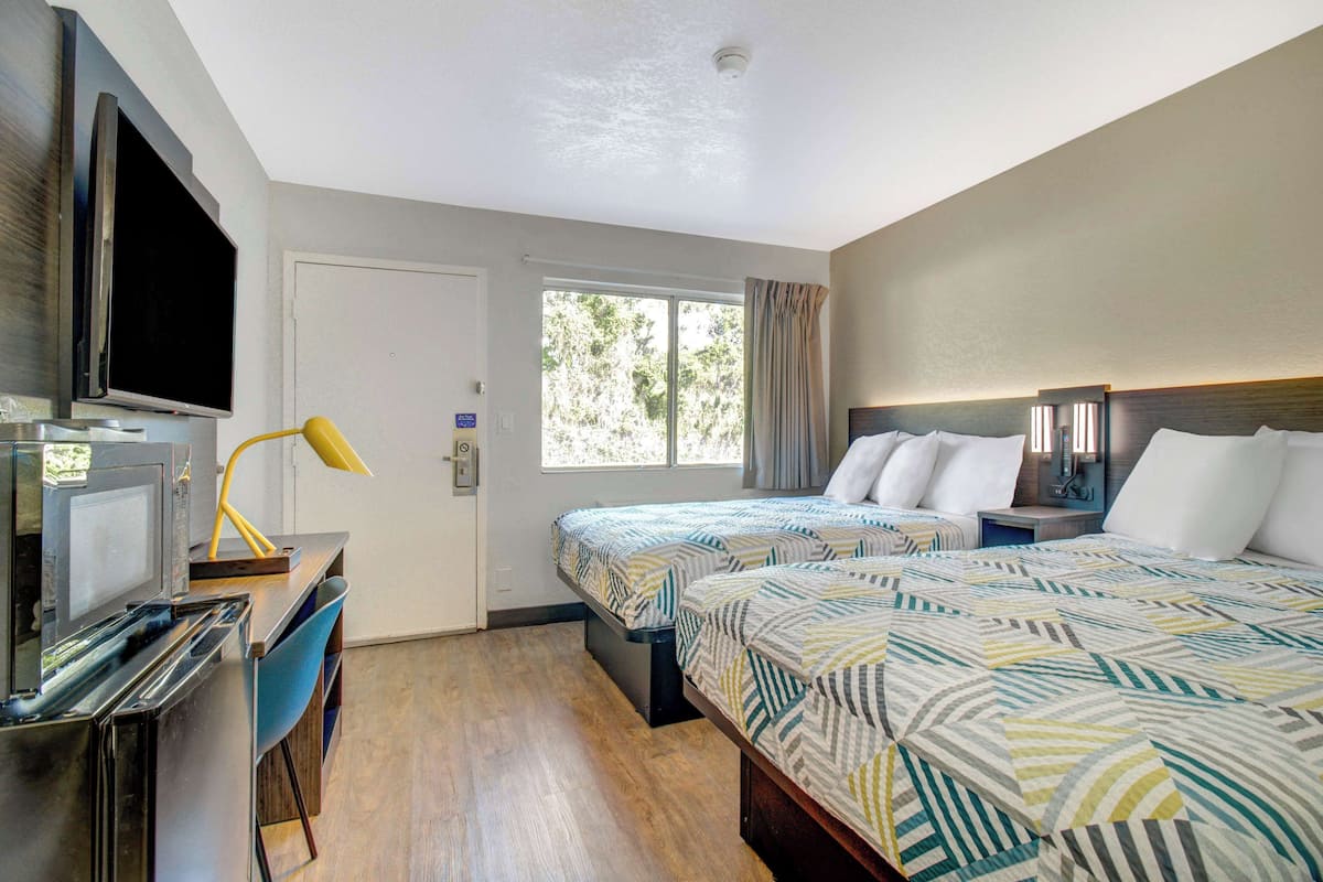 A hotel room with two beds, a desk with a computer and a lamp, a TV, and a window with curtains.