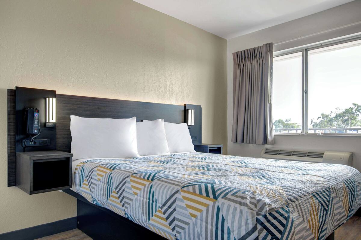 A hotel room with a bed, a nightstand with a phone, a wall-mounted lamp, and a window with curtains.