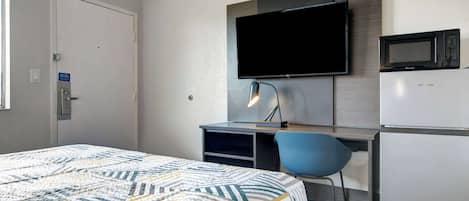 A hotel room with a bed, a desk with a chair, a flat-screen TV, a microwave, and a refrigerator.