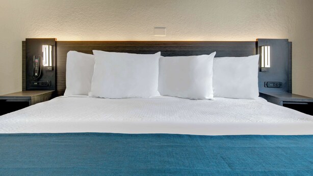 A neatly made bed with white linens and a blue throw blanket, flanked by two bedside tables with lamps. - Motel 6 Venice, FL (Venice)