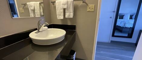 Un baño moderno con un lavamanos blanco, encimera negra y un espejo encima. Hay dos toallas blancas colgadas en un perchero.