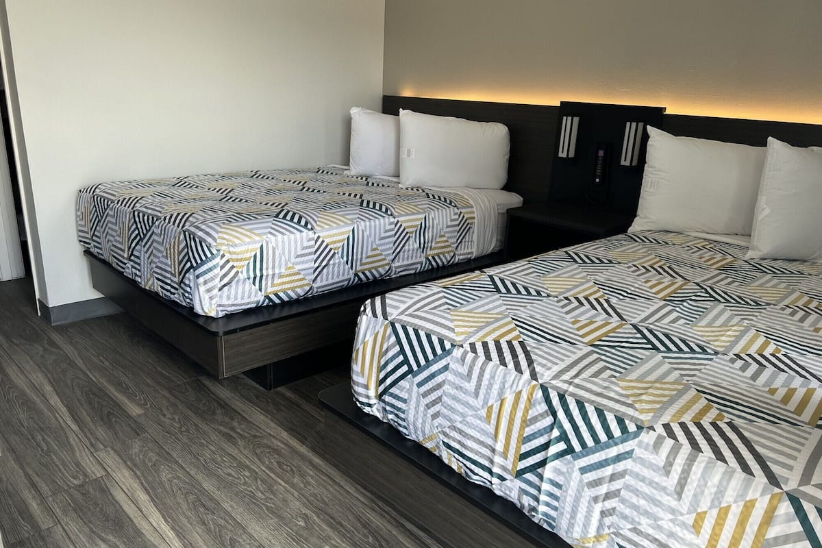 Dos camas individuales con ropa de cama de estampado geométrico, suelo de madera oscura y un panel de control en la pared.