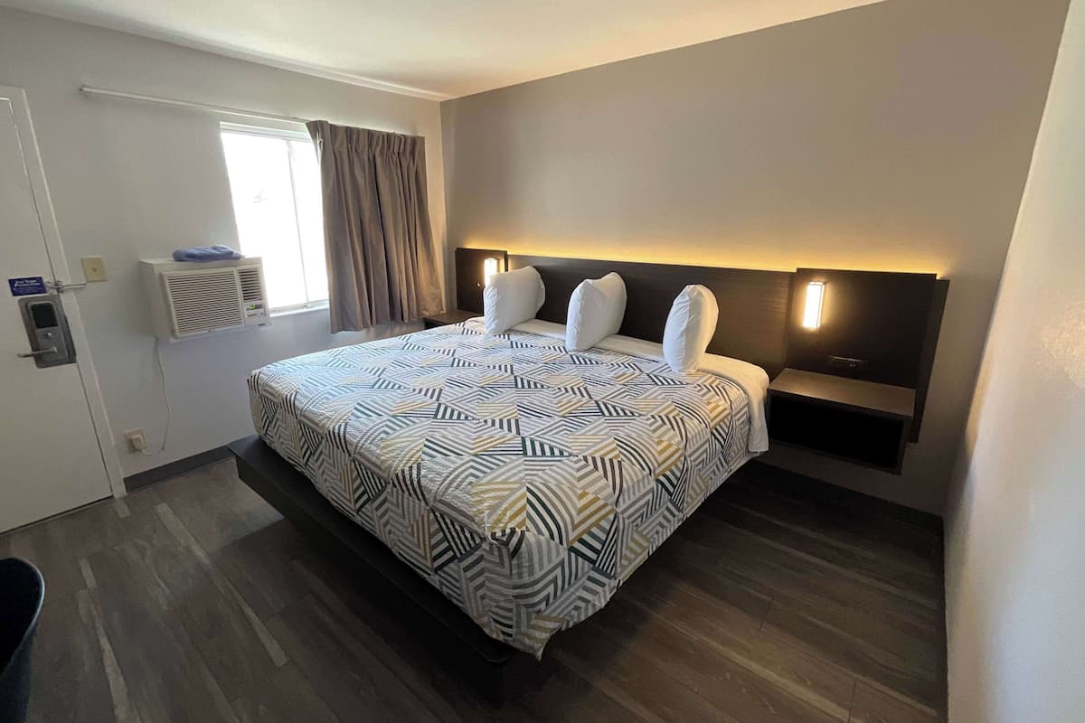 Habitación de hotel con una cama grande, una mesita de noche, aire acondicionado de pared, una ventana con cortinas y una puerta con un cartel.