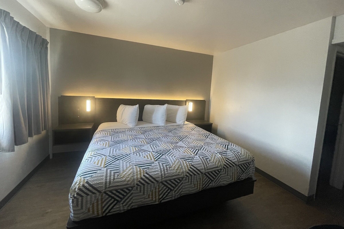 Una habitación de hotel con cama, mesita de noche, lámpara de pared y luz de techo.