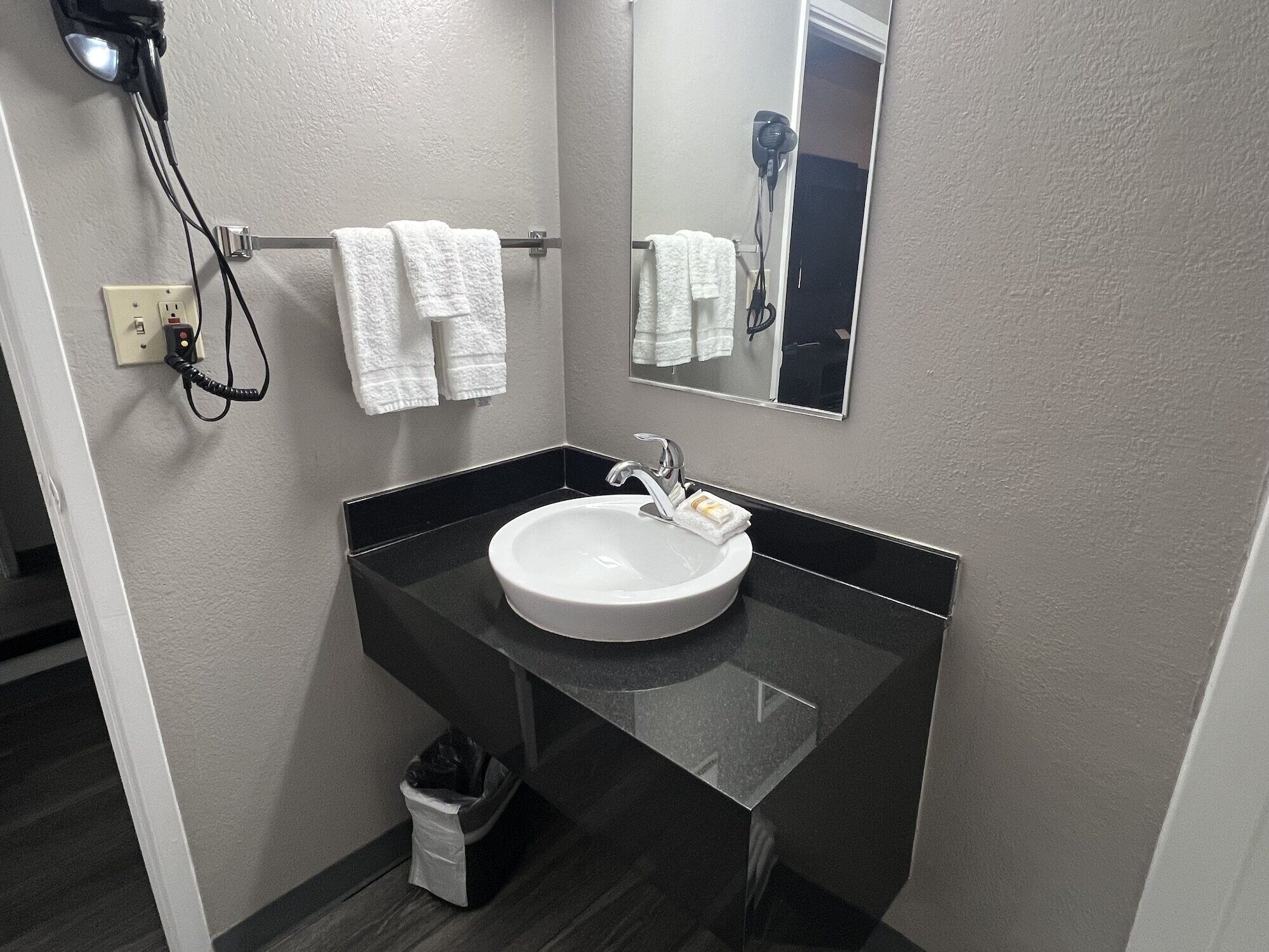 A bathroom with a black countertop, a white sink, and a mirror. There are two white towels hanging on a rack and a hairdryer on the wall.
