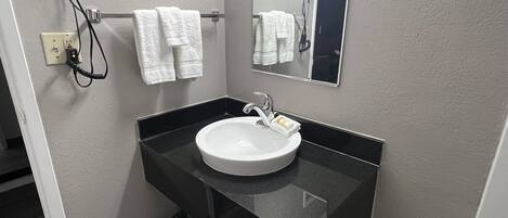 A bathroom with a black countertop, a white sink, and a mirror. There are two white towels hanging on a rack and a hairdryer on the wall.