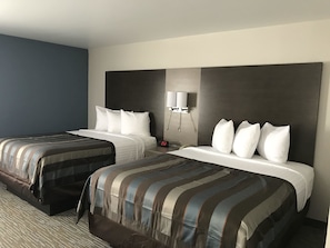 A hotel room with two beds, each with white linens and striped bedding. There is a nightstand with a clock and a wall-mounted lamp.