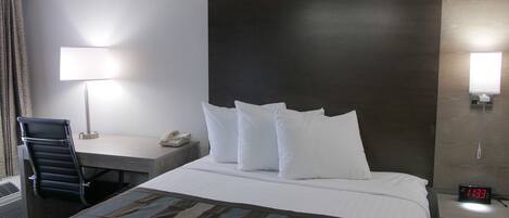 Habitación de hotel con cama, escritorio con lámpara, silla, teléfono y reloj digital.