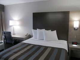 Habitación de hotel con cama, escritorio con lámpara, silla, teléfono y reloj digital.