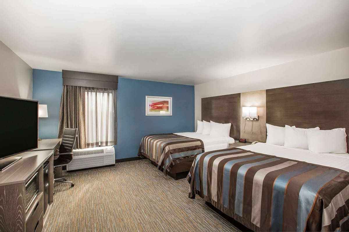 A hotel room with two beds, a desk with a TV, a chair, a window with curtains, and a wall-mounted light.