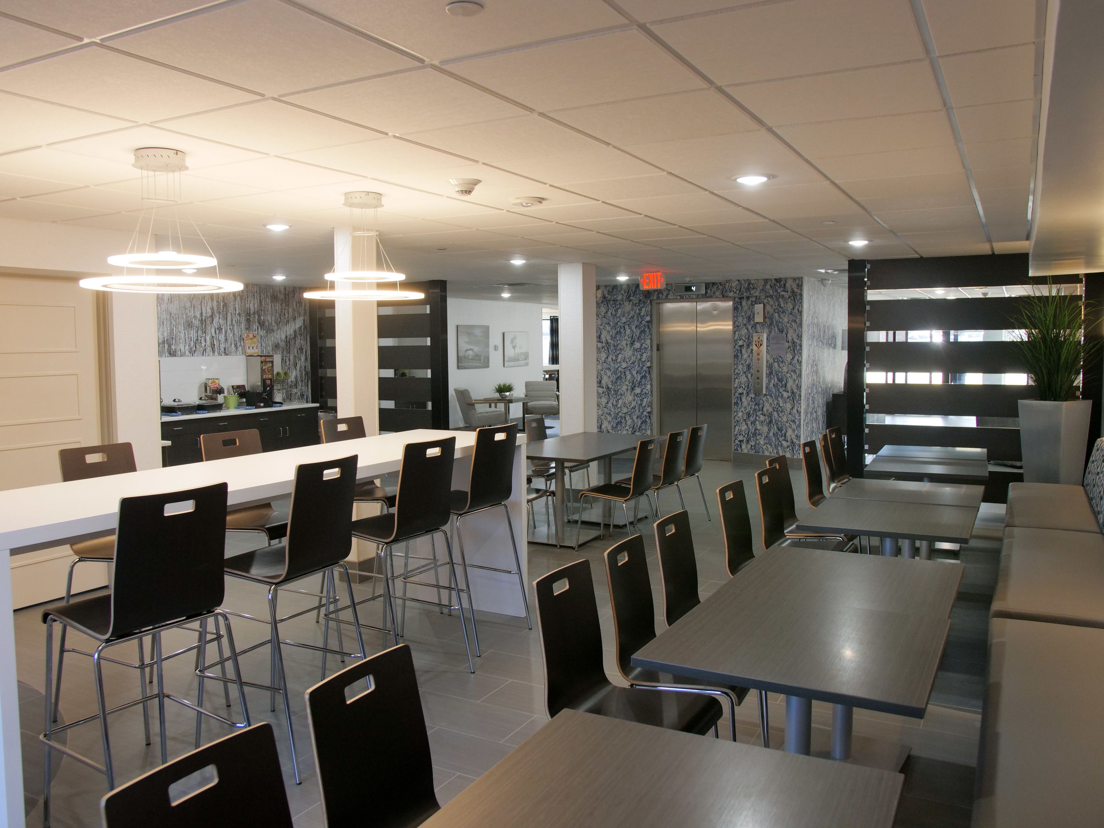 A modern dining area with multiple tables and chairs, a kitchen in the background, and a staircase leading to an upper level.