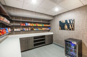 Una kitchenette con estantes llenos de refrigerios, un refrigerador y un cuadro en la pared.