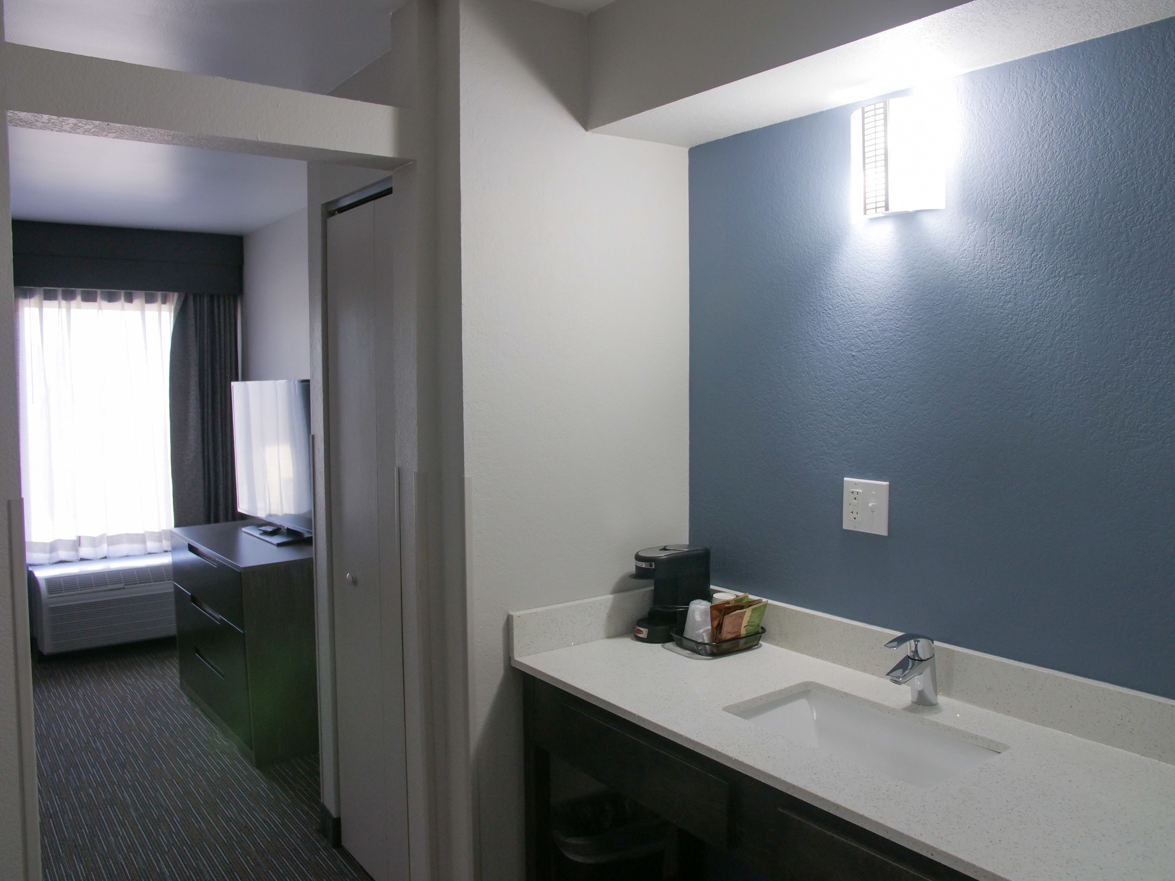 A hotel bathroom with a white sink, a mirror, and a towel rack. There is a window with curtains and a bed visible in the adjacent room.