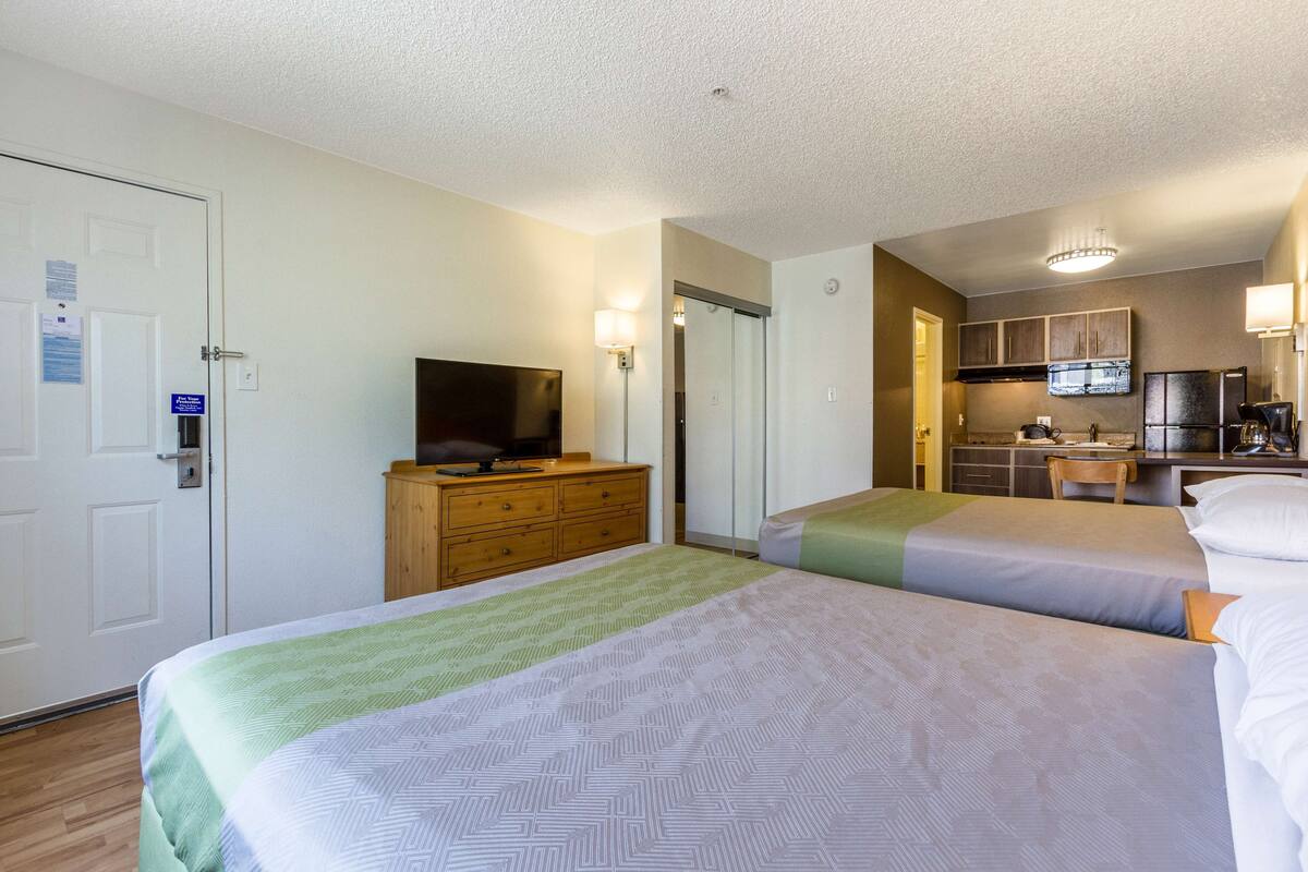 A hotel room with two beds, a TV on a dresser, a kitchenette, and a door with a sign.